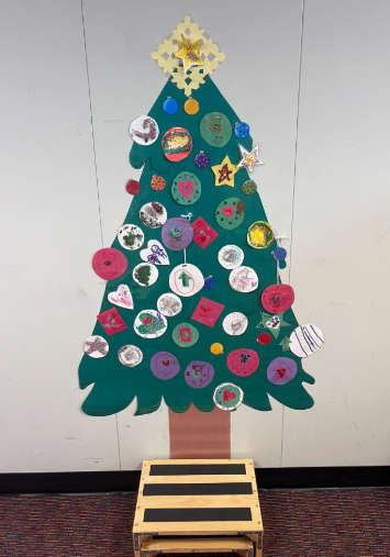A construction paper Christmas tree hung on a wall with a variety of construction paper ornaments all over it. 