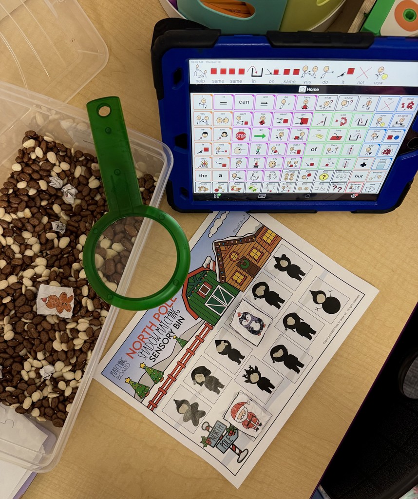 Image from above of a sensory bin with beans and a magnifying glass, a matching worksheet on the table with it, and a communication device to the side. 