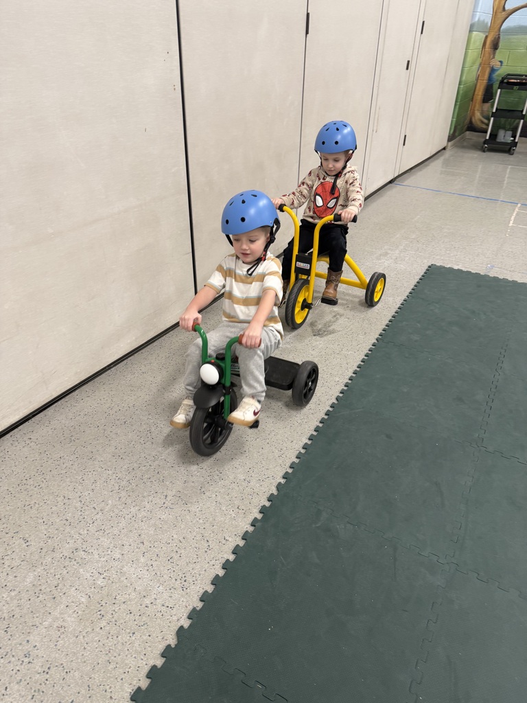 Two preschool children wearing helmets ride tricycles indoors, one following closely behind the other.