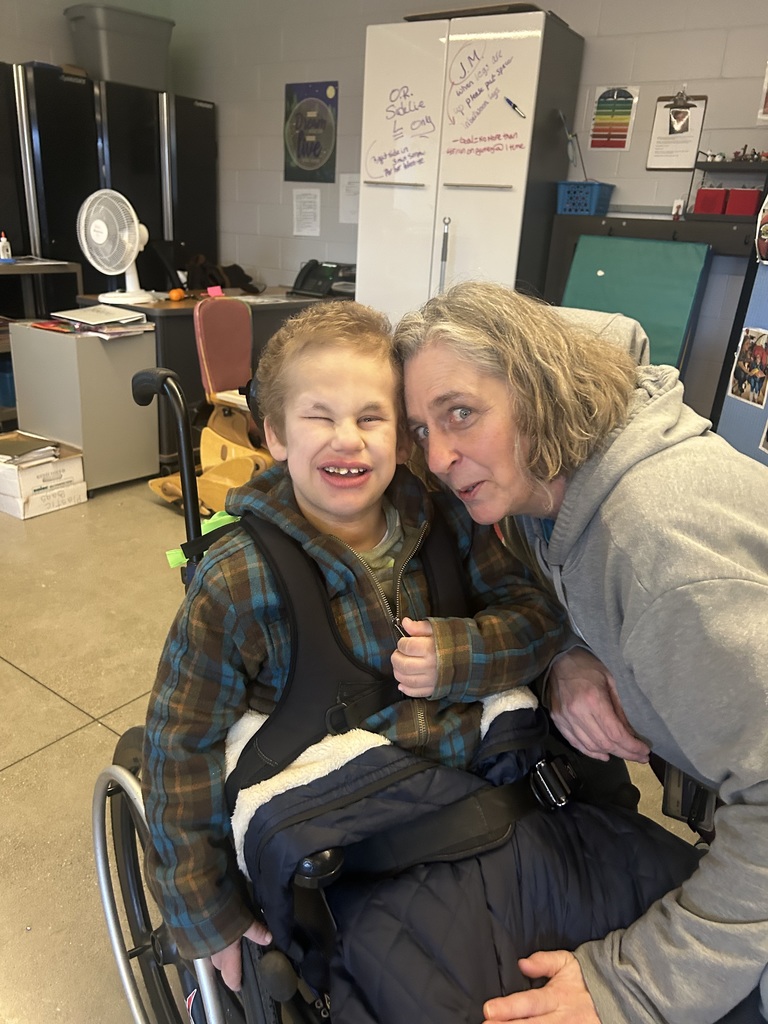 A woman being goofy, making a young student in a wheel chair smile.