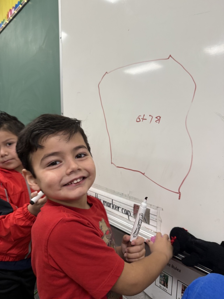 Smiling young child holding a dry-erase marker at a classroom whiteboard with a simple drawing and the numbers “6 7.”
