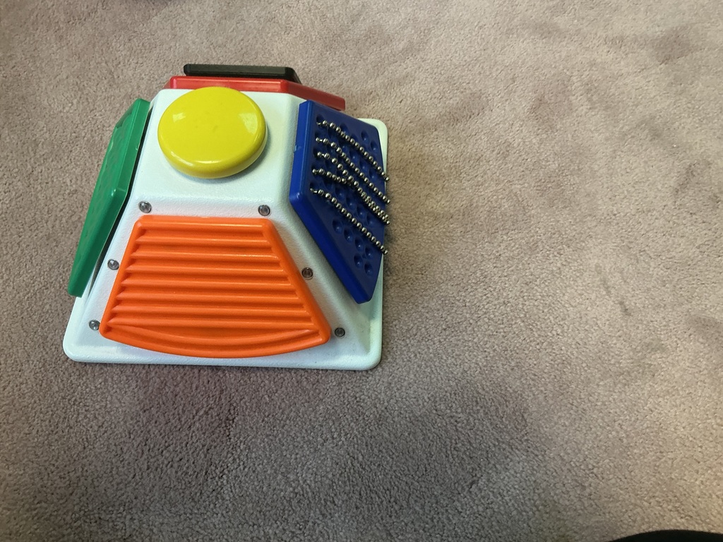 A tacticle communication tool that is cube shaped with different colored and textured buttons on each side. 