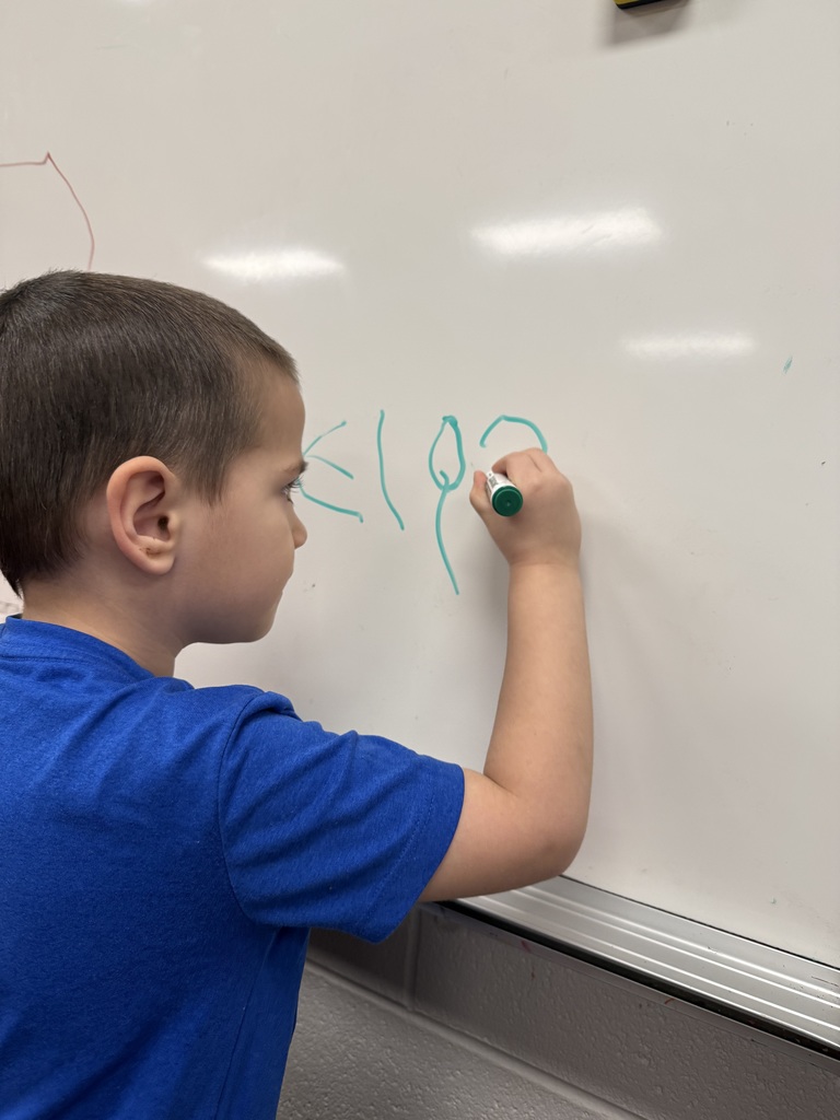 A preschool-aged child writing letters of their name on a whiteboard with a green marker.