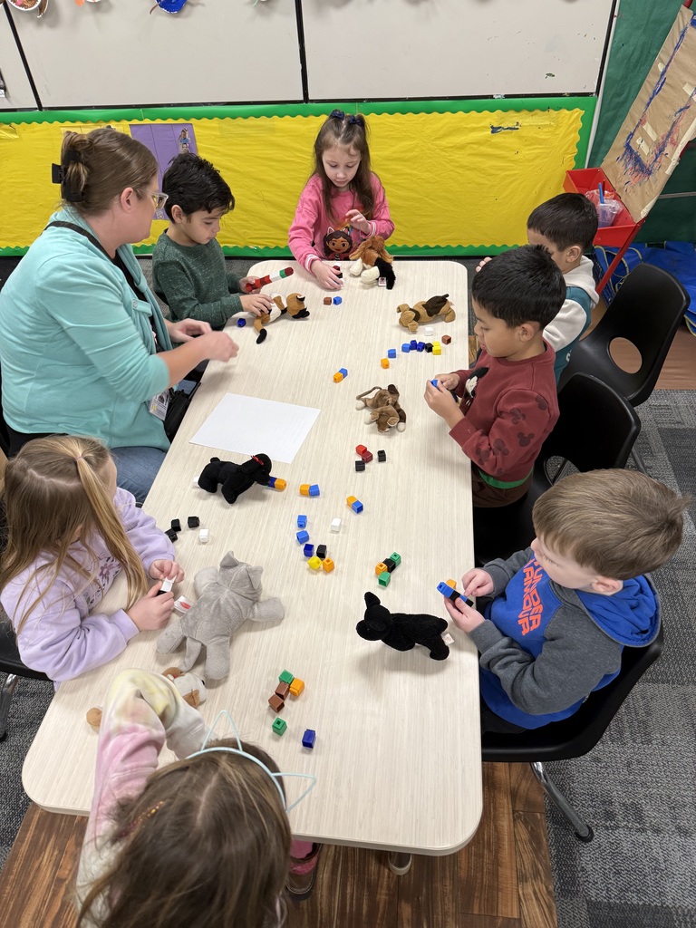 Preschool children sit around a table measuring toy pets with Unifix cubes while a teacher supports the activity.