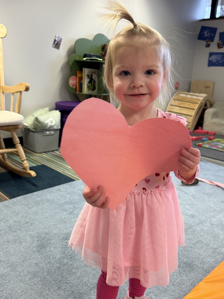 Toddler in a pink dress holding a large pink paper heart while standing in an Early Head Start classroom.