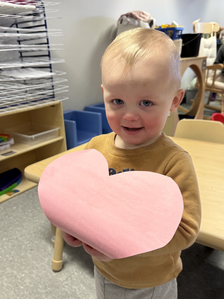 Toddler holding a pink paper heart and smiling while standing near a table in an Early Head Start classroom.
