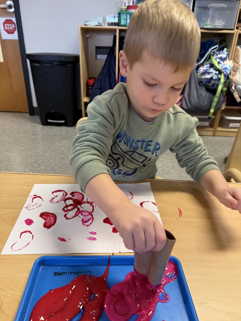 Toddler using a cardboard tube to stamp red and pink paint onto paper during an open-ended art activity in an Early Head Start classroom.