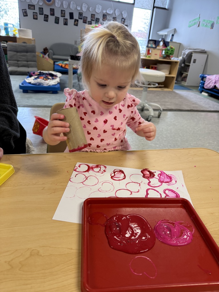 Toddler using a cardboard tube to stamp red and pink paint onto paper during an open-ended art activity in an Early Head Start classroom.