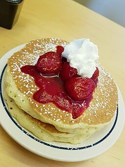Pancakes on a plate, topped with strawberries and whip cream.