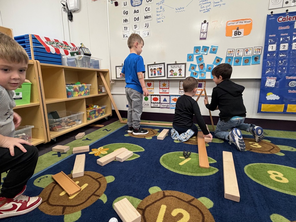 Several preschool students work together on the carpet to build a long wooden ramp system, watching a ball roll down as they test distance, speed, and stability.