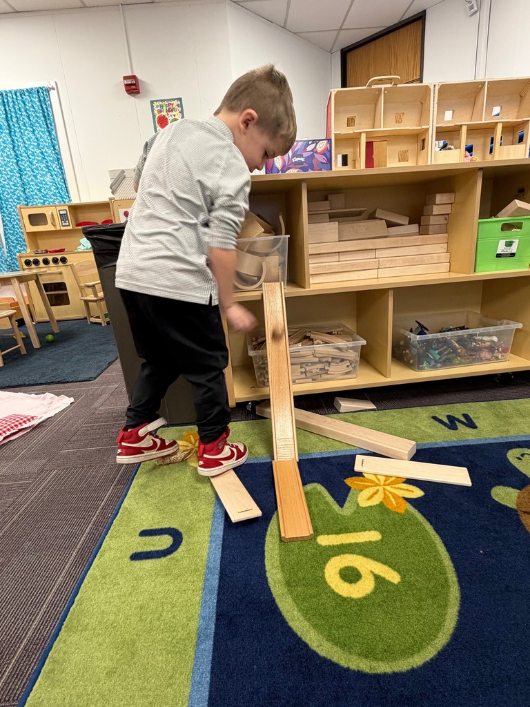 A preschool student stands at a low shelf, arranging wooden planks into a ramp and adjusting the angle to roll a ball downward.