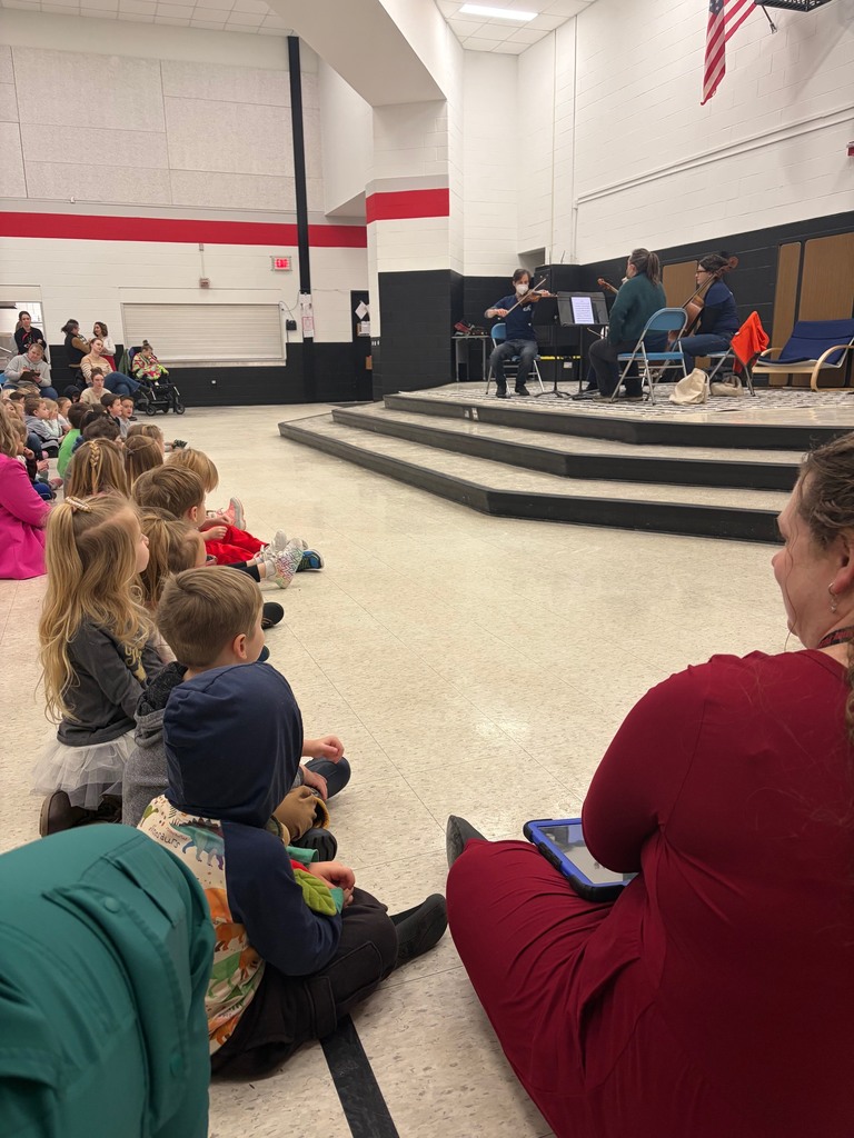 Preschool students sit together on the floor, watching and listening to an orchestra perform during a school assembly.