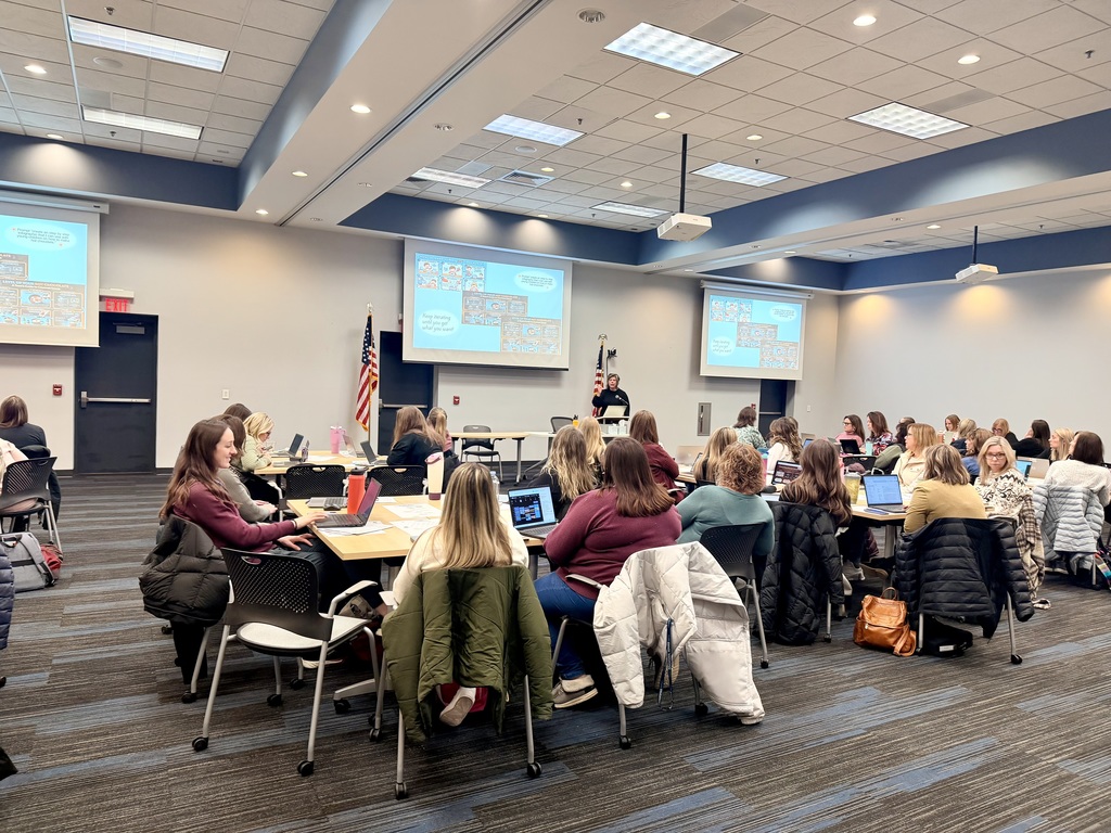 Room with a large group of folks seated around tables and viewing a presentation on 3 large screens. 