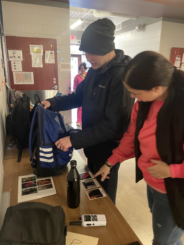 Teacher is pointing to visual schedule, while students is unzipping backpack.