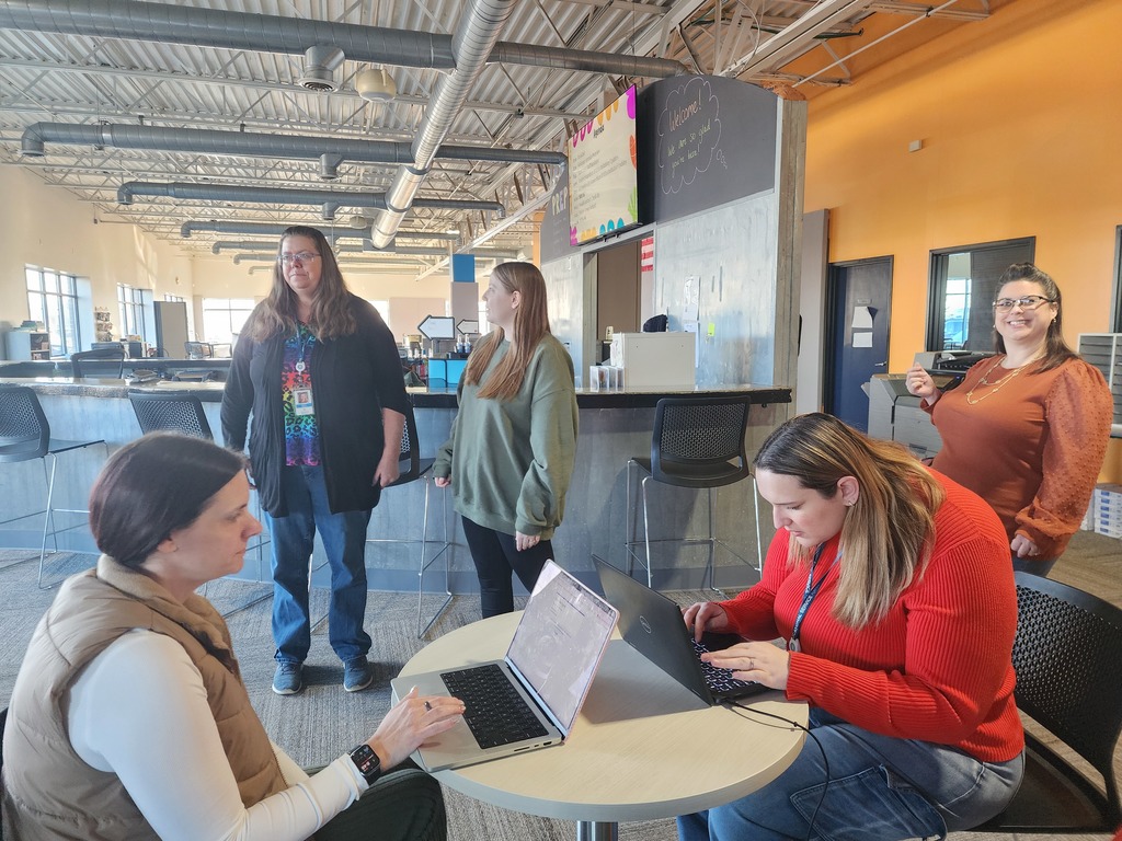 Women standing and waiting while two women sit at a table working on their laptops.