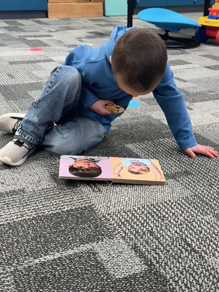 A young child sits on the classroom carpet looking at a small board book while holding a breakfast snack in his hand.