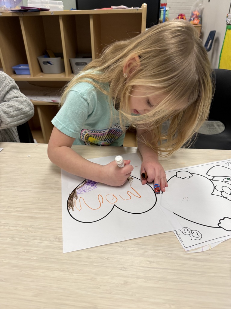 A young child sits at a table coloring and writing the word “mom” from memory on a heart-shaped worksheet.