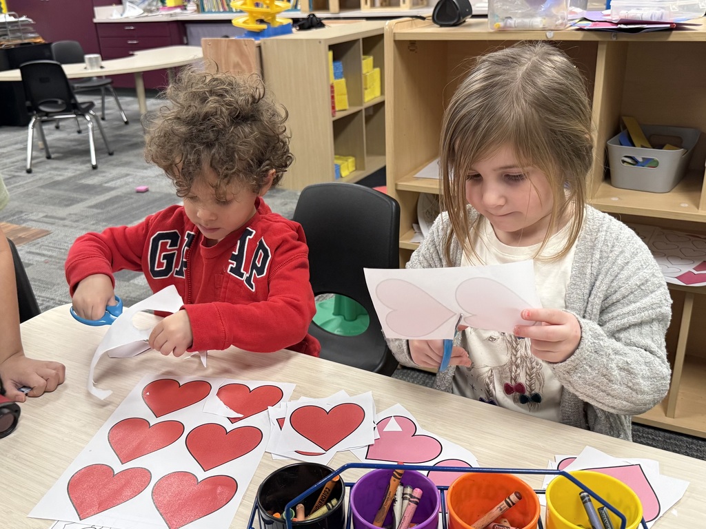Two young children sit at a classroom table using scissors to cut out paper hearts.