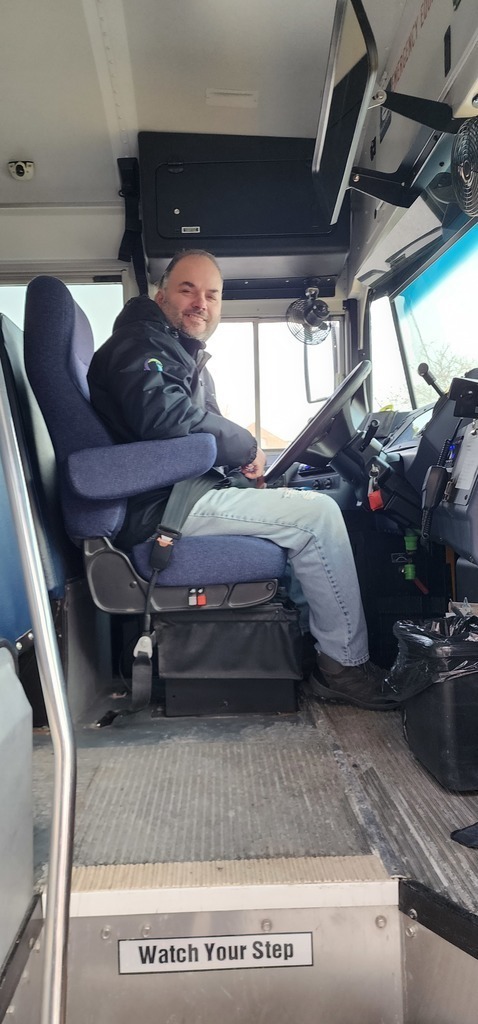 An adult male sitting in the driver seat of a bus.
