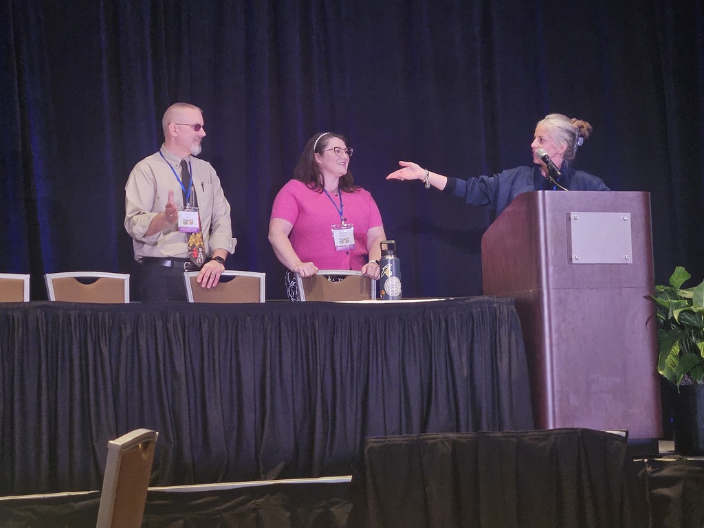 Group of people on a stage one lady is standing behind a podium with her arm outstretched towards the other 2 people 