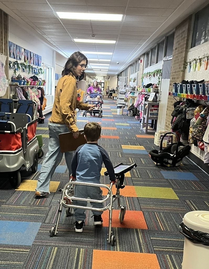 Student with a walker and AAC device mounted on the walker is walking down the hallway with a teacher. 
