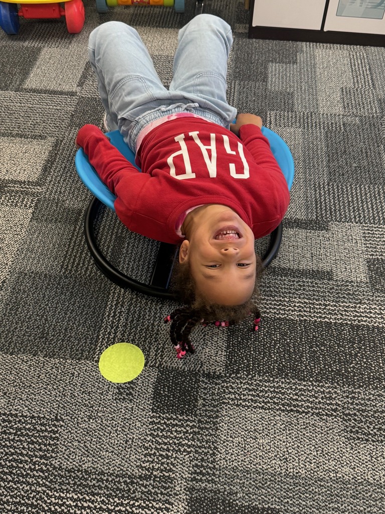 Preschool child smiling upside down while lying on a blue wobble seat in a classroom, with a green floor marker nearby.