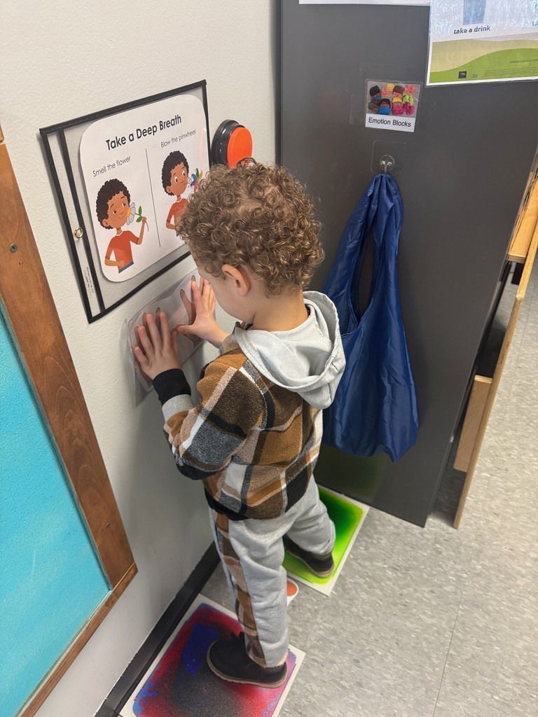 Preschool children standing in the classroom Big Breath area, pressing their hands to the wall beneath a “Take a Deep Breath” poster while standing on sensory floor spots.