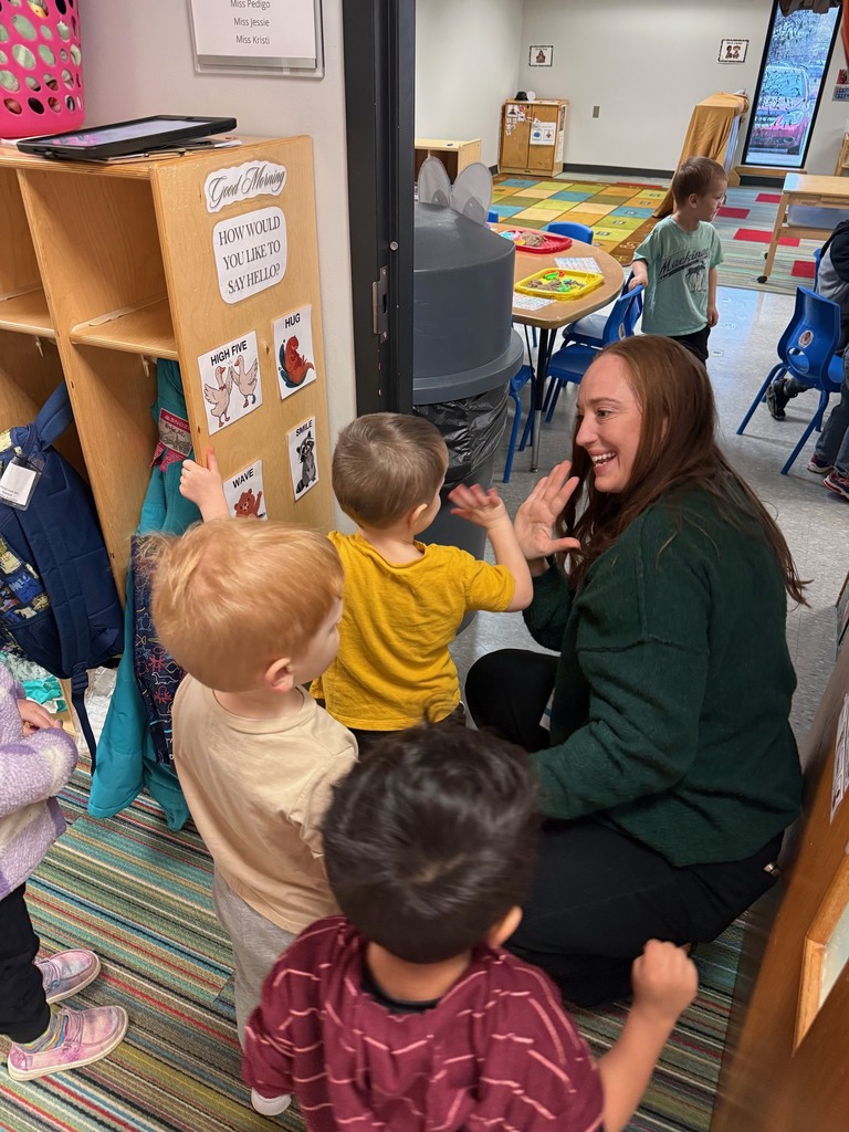 A teacher kneels at the classroom entrance smiling as children choose how to say hello using a greeting board with options: high five, hug, wave, or smile.