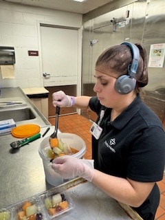 A food service worker wearing a hairnet, gloves, and over-the-ear headphones scoops diced fruit from a large white bucket into clear plastic containers in a commercial kitchen.