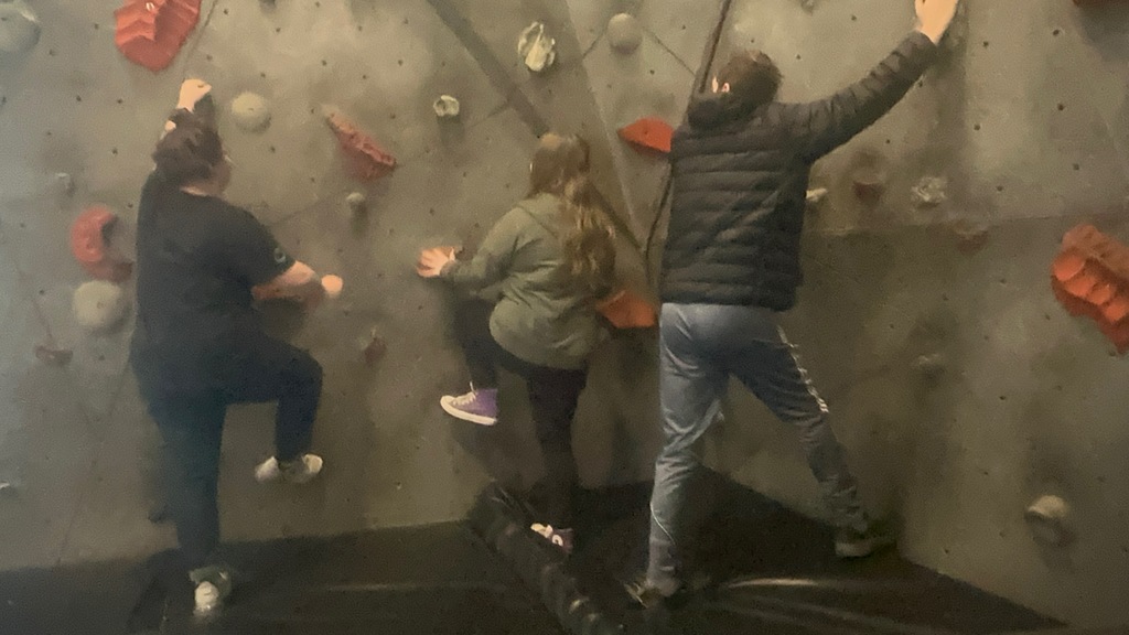 Three individuals scale an indoor rock climbing wall from behind, using various colored handholds above a padded black floor.