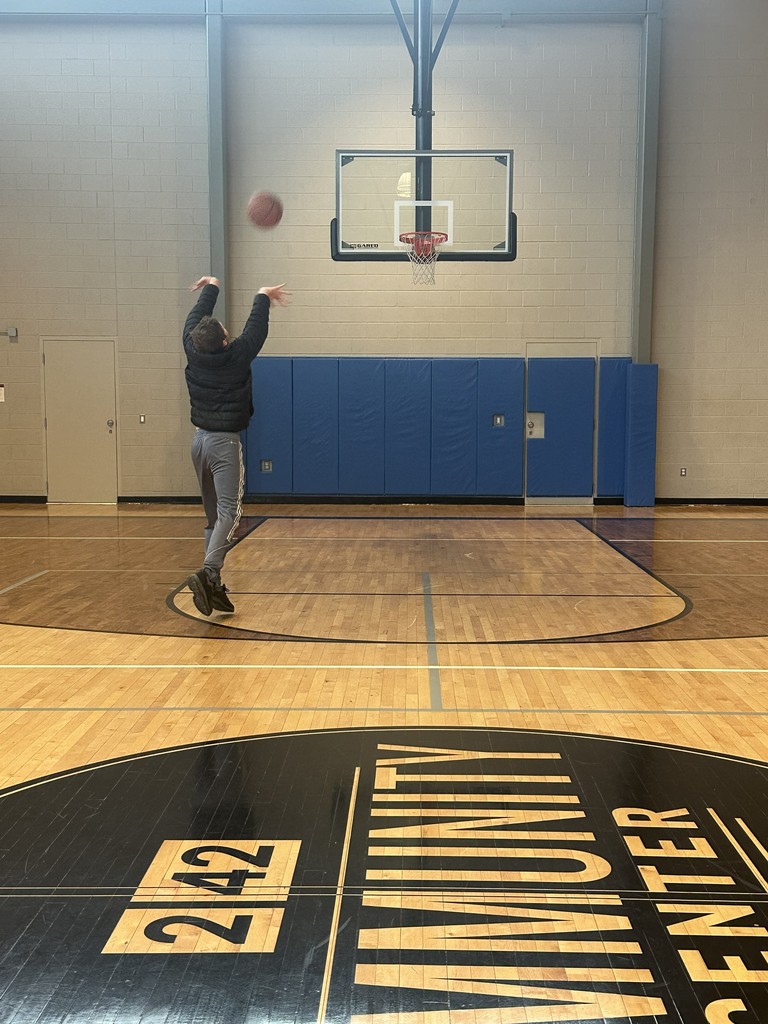 A person in mid-air shoots a basketball toward a hoop in an indoor gymnasium. The court floor in the foreground features a large black and tan logo for the "242 Community Center