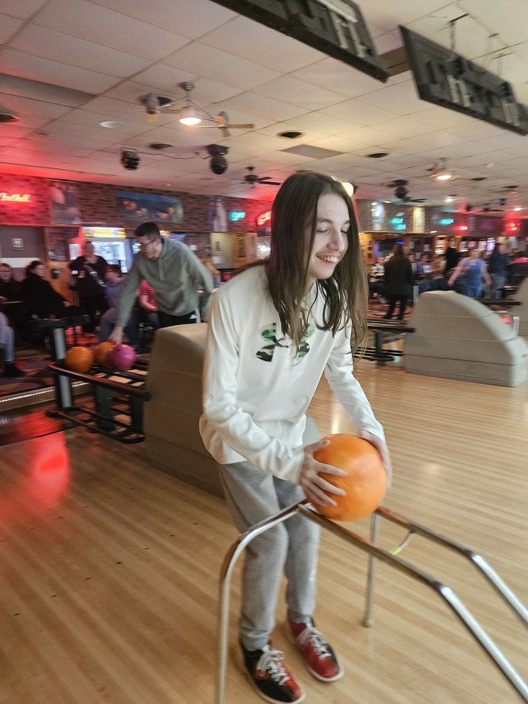 A young person with long hair and a white Under Armour long-sleeved shirt is seen smiling while positioning an orange ball on a ramp.