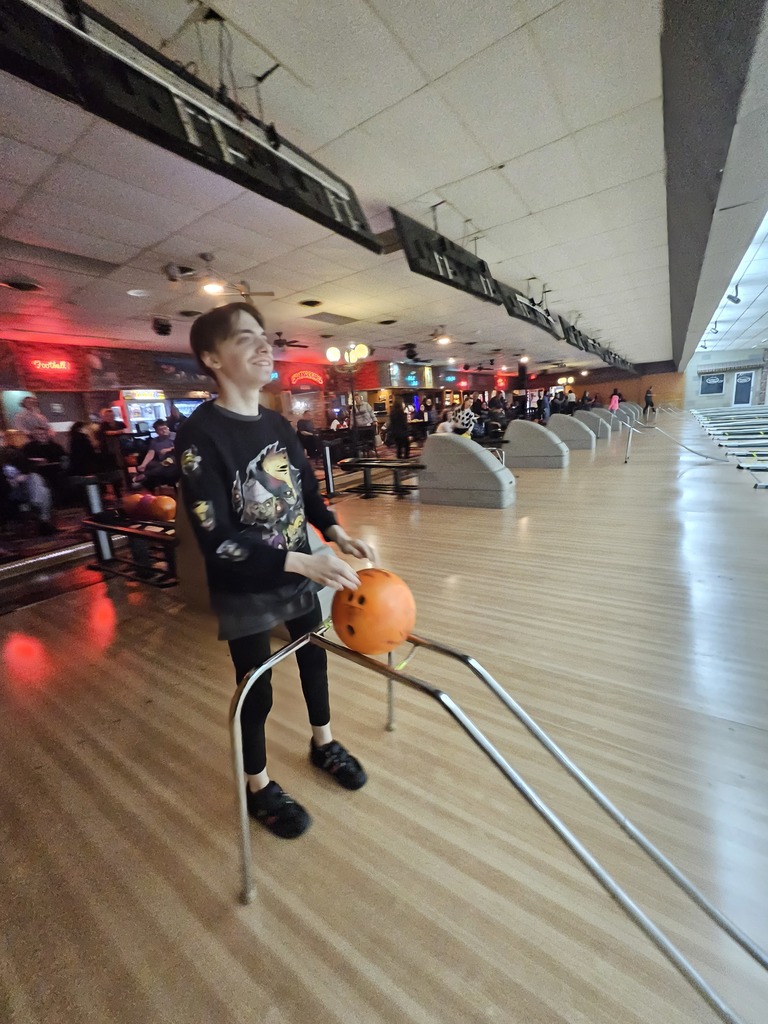 One young man in a sage green hoodie and black athletic pants is shown mid-motion, releasing an orange bowling ball down a lane.