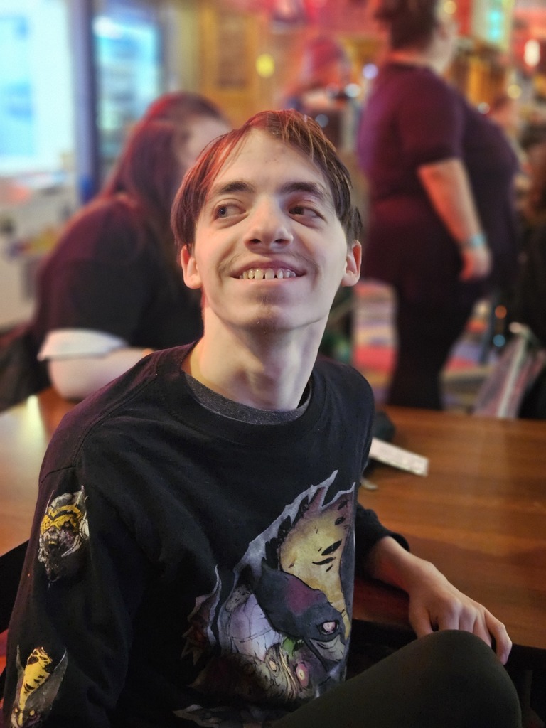 Another portrait features a young man in a black long-sleeved graphic t-shirt looking upward with a bright smile while seated at the table.