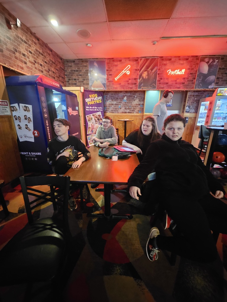 Four individuals are seated or standing around a dark wooden table. A young man in a black graphic t-shirt sits on the left, while a young man in a grey hoodie and a woman in a black shirt are seated behind the table. In the foreground, another individual in a black fleece jacket looks directly at the camera.