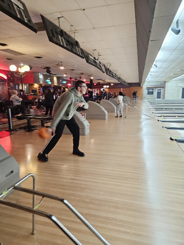 image shows a young man in a sage green hoodie and black athletic pants mid-motion as he releases an orange bowling ball down a lane.