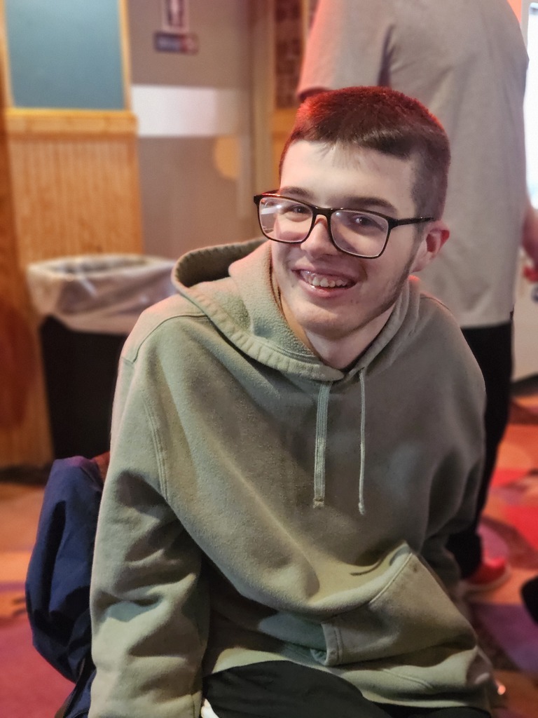 A young man with glasses and a short haircut is shown in a medium close-up, smiling warmly toward someone off-camera.