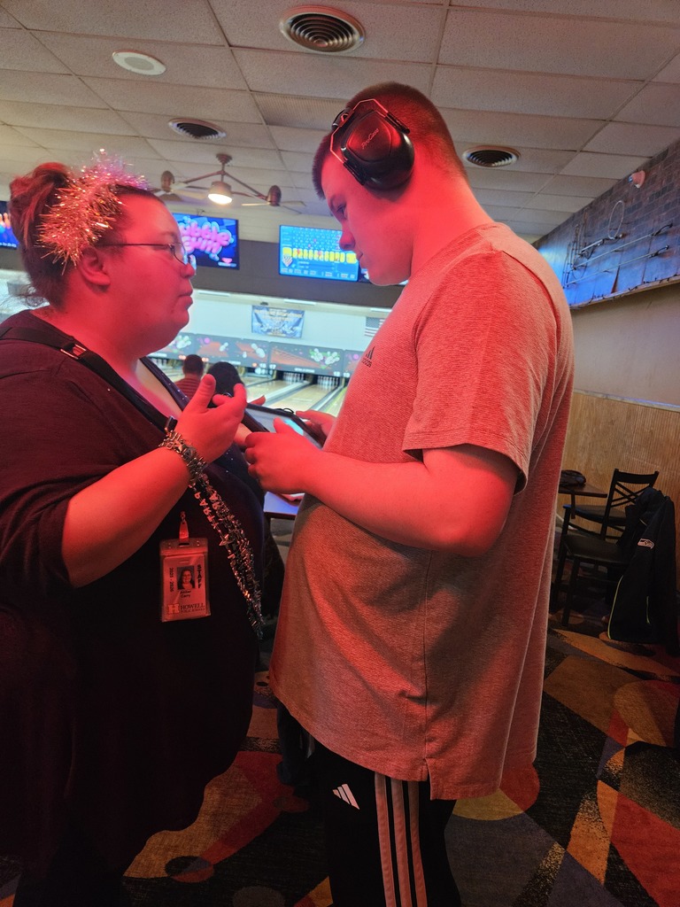 On the left, a woman with glasses and festive tinsel in her hair wears a black top and a staff lanyard. She is gesturing with her hands while speaking to a young man on the right. The young man, wearing a grey t-shirt, black athletic pants, and large noise-canceling headphones, is looking down at a tablet or device they are holding together.