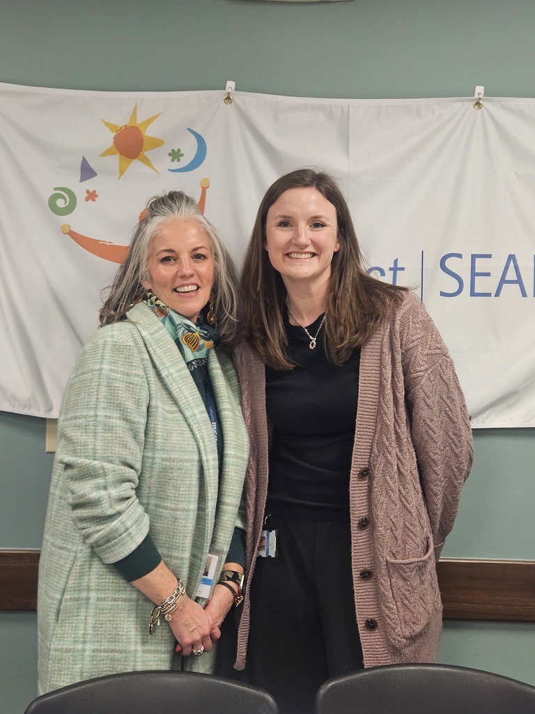 Two women stand side-by-side, smiling in front of a white banner. On the left, a woman with long silver hair wears a light green plaid coat and a patterned scarf. To her right, a woman with long brown hair wears a mauve cable-knit cardigan over a black top. The banner behind them features a colorful logo with a sun and crescent moon, along with the visible text "Project | SEARCH."