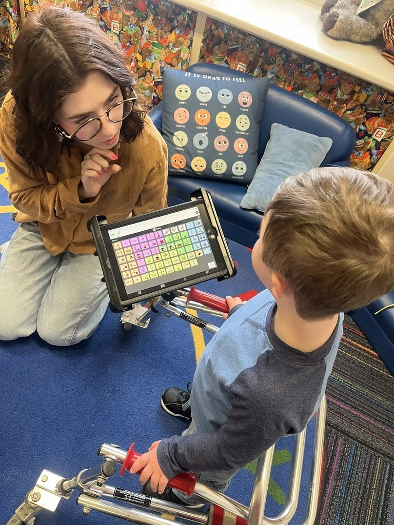 Overhead view of a student with a walker and an AAC device mounted on the walker and a teacher signing and talking with the student. 