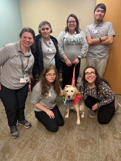 An indoor, eye-level shot captures six people and a dog in a small room. In the back row, four people are standing from left to right: a woman with shoulder-length dark brown hair wearing a gray polo and black pants; a person with short brown hair wearing headphones, a gray polo, and a black jacket; a woman with long dark brown hair and glasses wearing a gray sweatshirt; and a man with short brown hair wearing a gray polo and light gray pants. In the front row, two people are kneeling on the patterned tan carpet: a woman on the left with long brown hair and glasses wearing a gray T-shirt and black pants, and a woman on the right with long dark brown hair and glasses wearing a floral print blouse and black pants. Between them sits a yellow Labrador Retriever wearing a rainbow-colored ruffled collar. The woman in the front row on the left has her hand on the dog's head. The woman in the back row with the sweatshirt holds the dog's red leash. The wall on the left is light blue, and the wall in the back is light gray. A light brown wooden door is on the right. Everyone in the group is smiling.