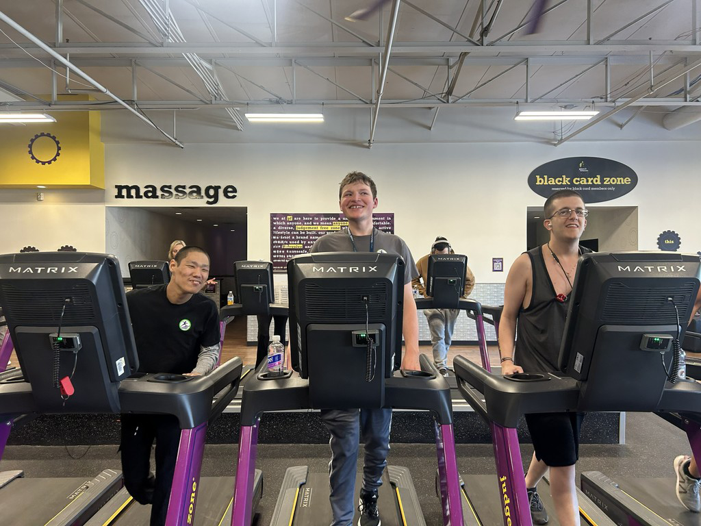 A wide shot captures three young men standing on Matrix treadmills in a Planet Fitness gym, all smiling at the camera. The gym features a "massage" area on the left and a "black card zone" on the right. The man on the left is wearing a black t-shirt, the man in the middle is in a grey t-shirt, and the man on the right is wearing a black tank top and glasses. In the background, other gym-goers are visible, including a person in a blue shirt. The gym's ceiling is exposed, with fluorescent lights and pipes.