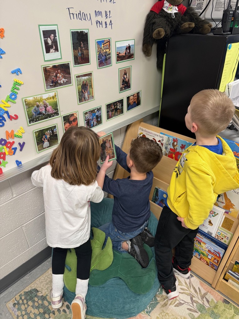 Three preschool children look at and hold family photos displayed on a classroom wall.