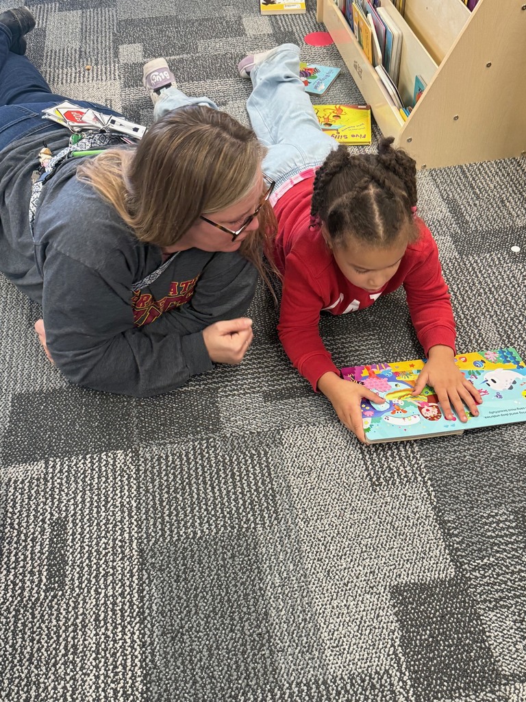 Teacher and preschool child lying on the classroom carpet reading a colorful board book together near a bookshelf.