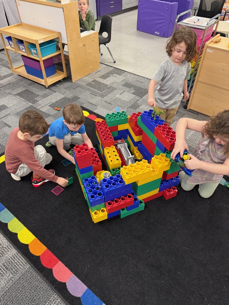 Four preschool children sit on a classroom rug building one large structure together with oversized colorful blocks.