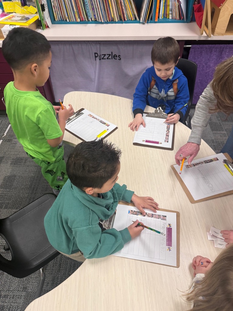 Preschool children stand and sit around a table using clipboards to sign in as they arrive at school.