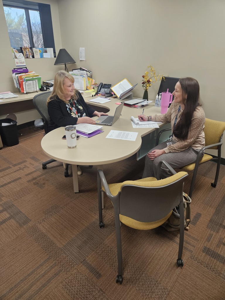 Two women are collaborating at a curved, light-colored wooden desk in a professional office setting. They are both smiling warmly at each other, creating an atmosphere of positive teamwork.