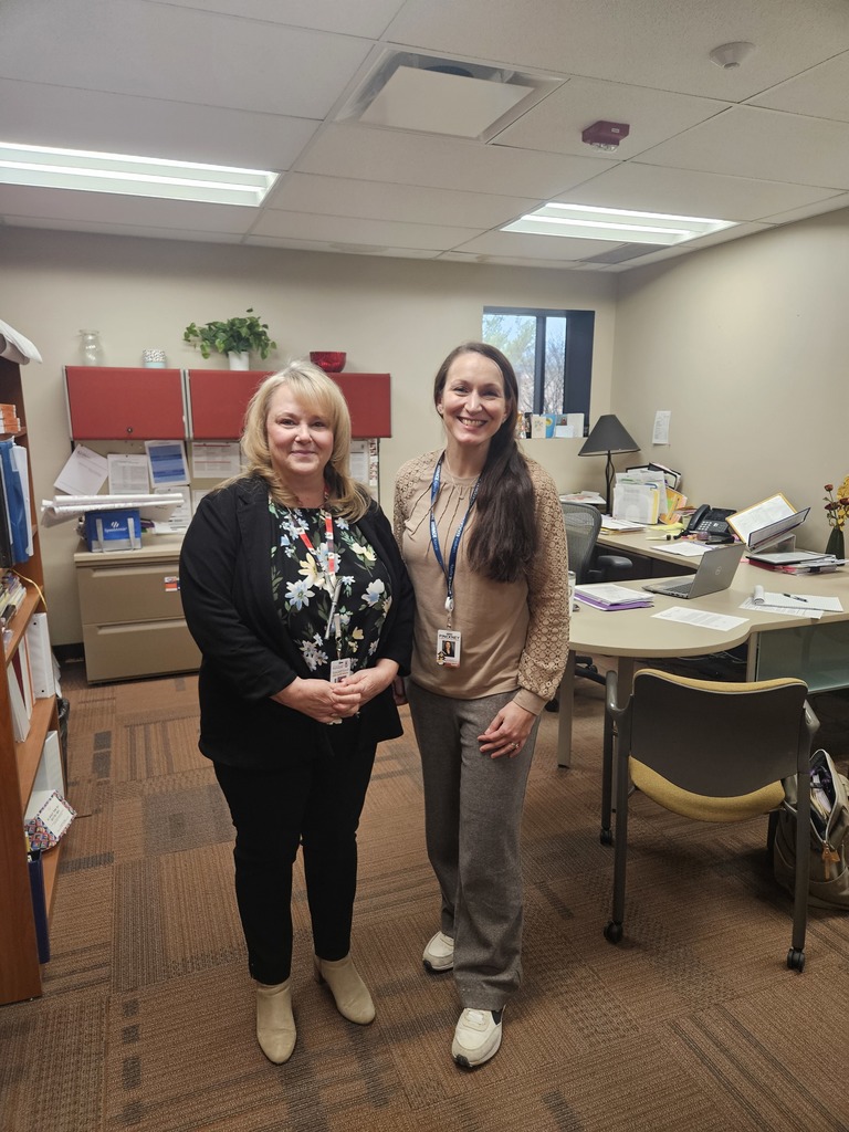 Two women stand side-by-side in the center of an office, smiling at the camera. They are dressed in professional attire and wearing identification lanyards. The background shows their workspace, including desks, a bookshelf, and a filing cabinet.