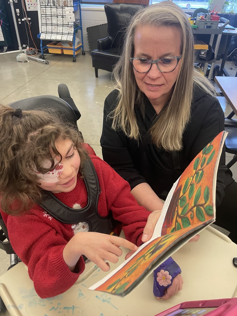 Student point to text in a book. Adult sitting next to her holding the book.