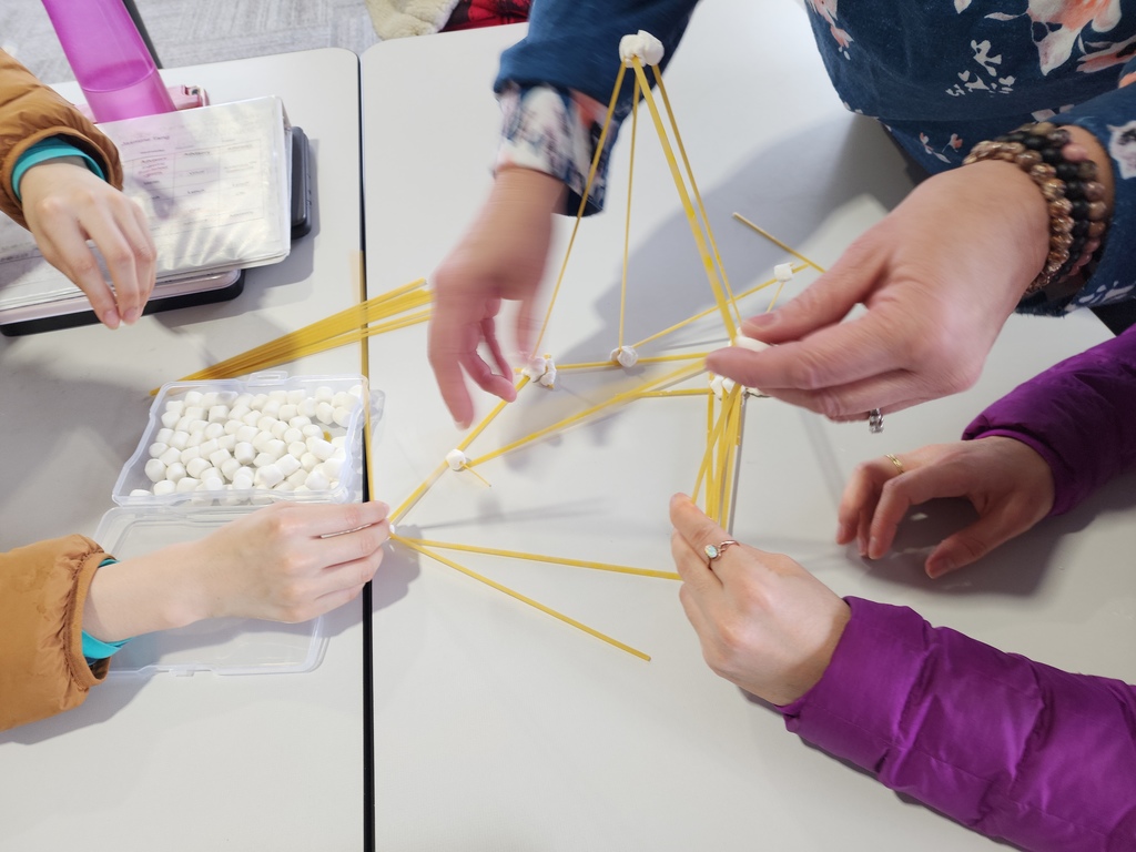 A close up of hands around a table building with spaghetti noodles and marshmellos.